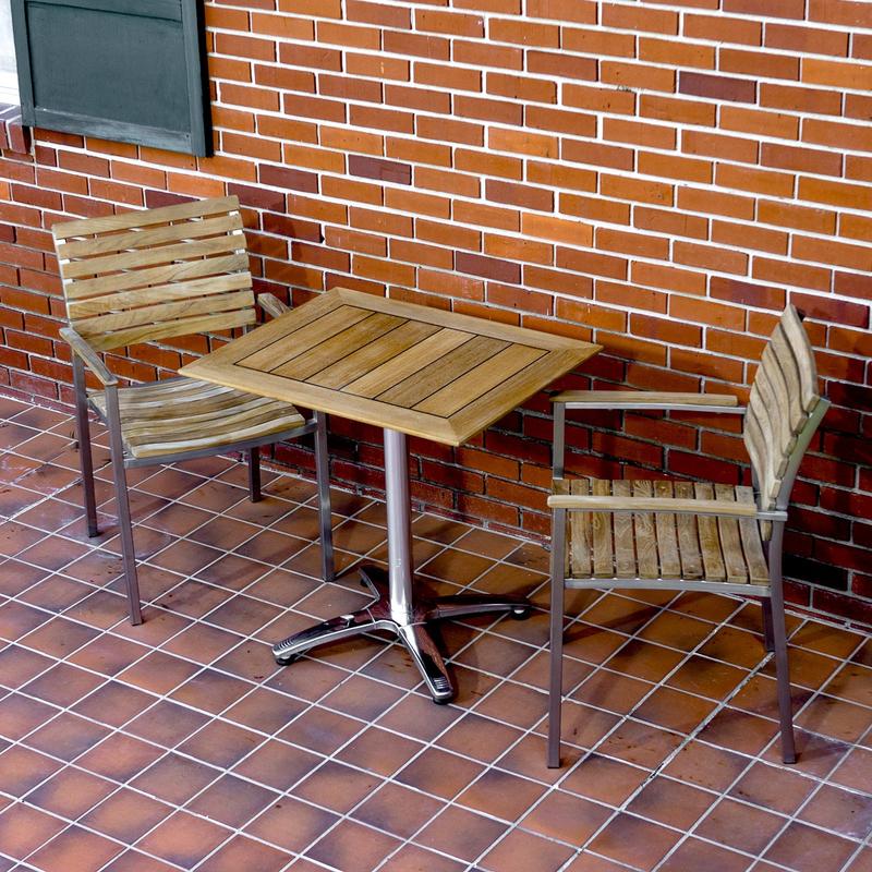 alt="70668 Veranda Vogue Bistro Set of teak and stainless steel rectangular table and 2 teak dining chairs on tiled outdoor patio against brick house and green window shutter"