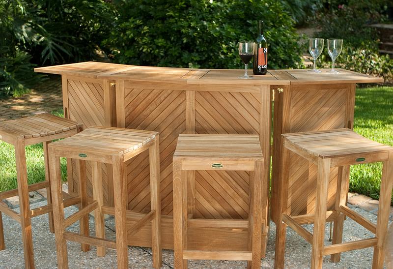 alt="17112 Somerset teak bar and stool five piece set showing three wine glasses and wine bottle on top with stone patio a grass area paver walkway and lush tropical plants in background"