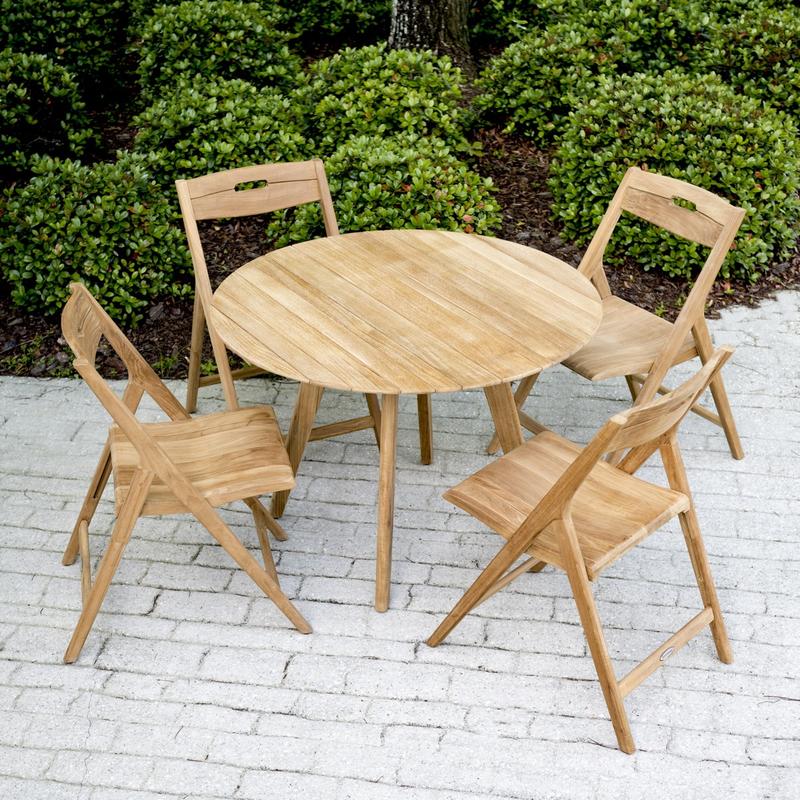 alt="70519 Surf Round Teak Dining Table Set of a 42 inch Round Teak Dining Table and 4 Surf folding chairs on white brick patio with shrubs and trees in background"
