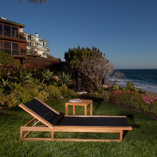 alt="16771 Maya Chaise Lounger in black Textilene mesh fabric and side table with magazine on a green lawn overlooking the ocean with two homes and flowering plants and trees in background "