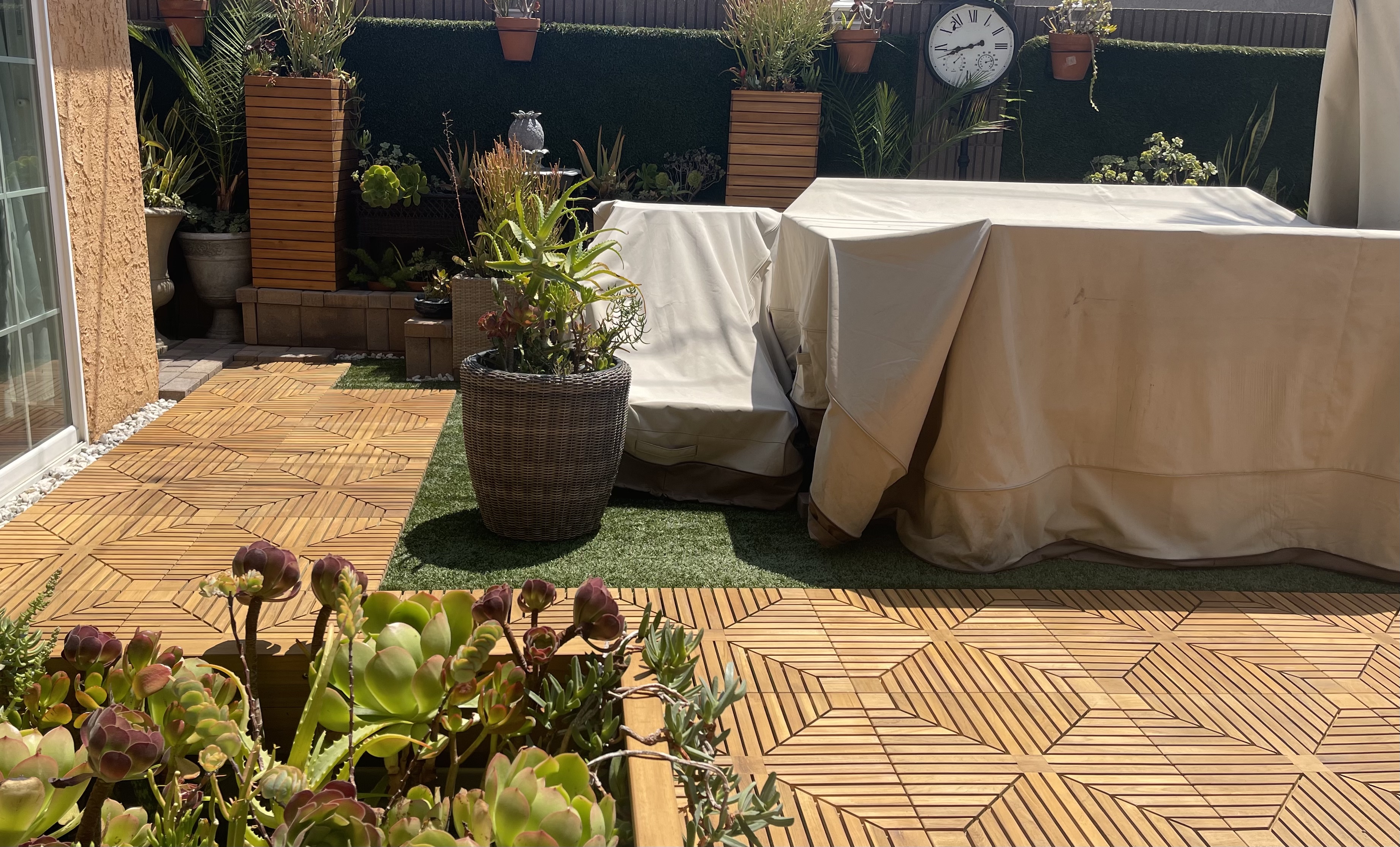 Beautiful Patio with teak flooring tiles with succulents in the foreground and covered outdoor furniture and planters and hedge in the background